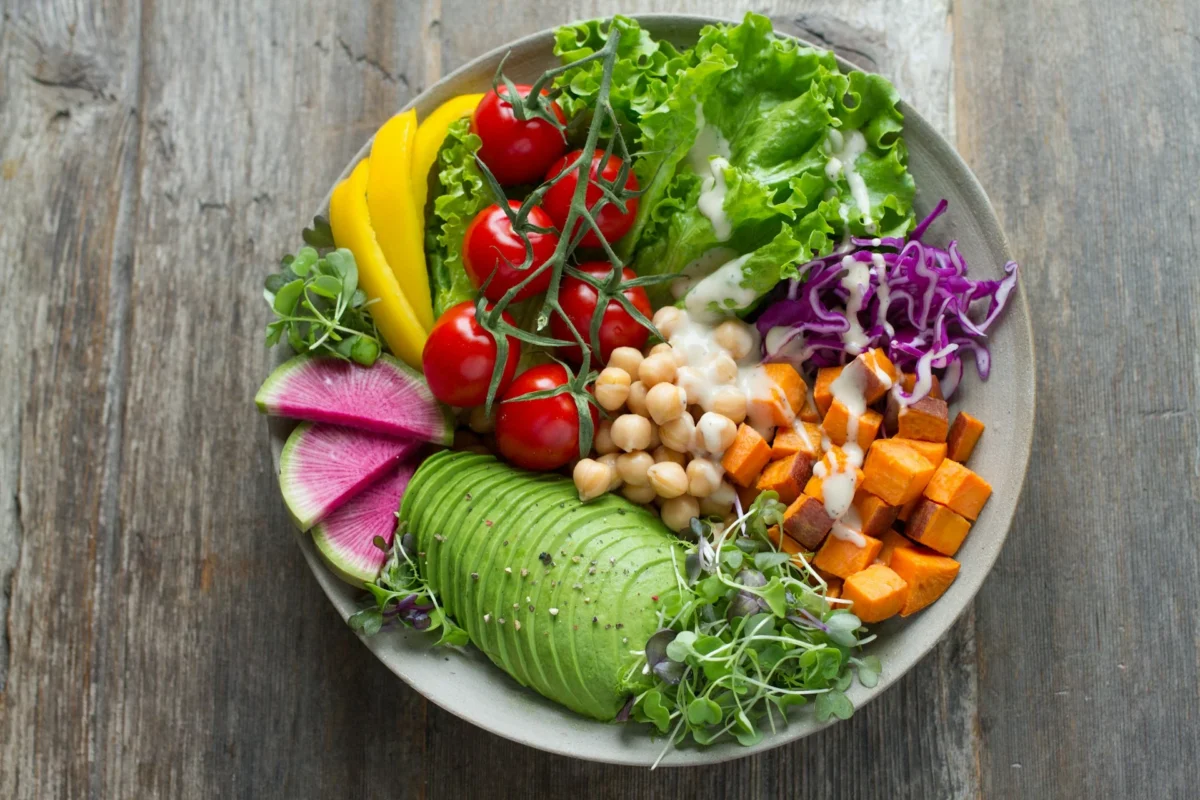 A colorful salad bowl with fresh avocado, cherry tomatoes, yellow bell peppers, lettuce, chickpeas, sweet potatoes, radishes, and microgreens, topped with a light dressing.