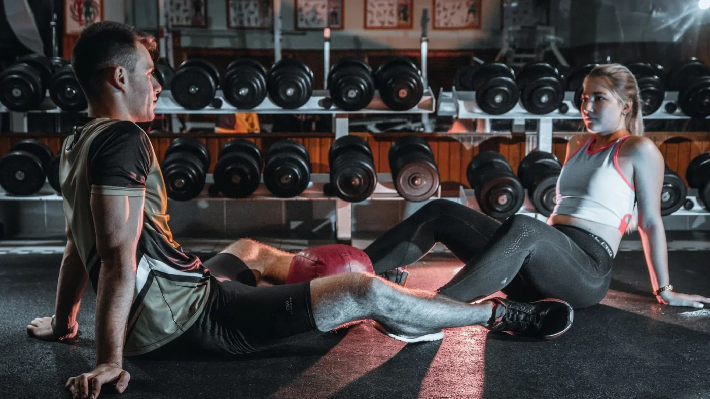 Two gym members resting on the floor in front of a rack of dumbbells between sets