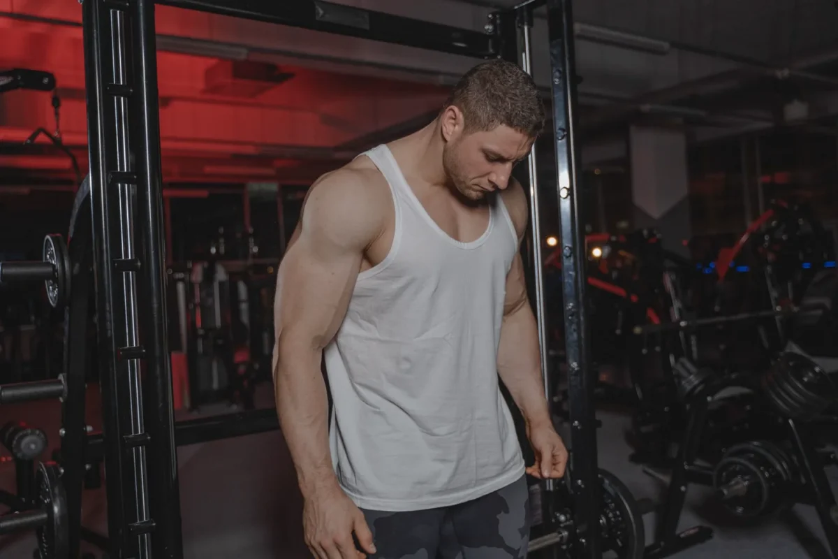 Muscular man in a white vest standing beside weight-training equipment in a dark gym