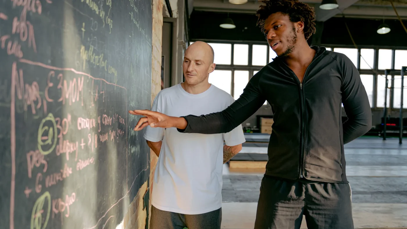 Two gym members discussing a training plan beside a blackboard